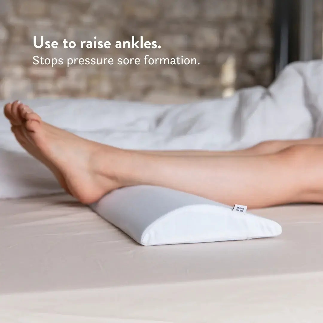 Person using a white elevation pillow to raise ankles on a bed.
