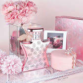 A pink perfume bottle with silver crystal-like circular embellishments, a round pink box, a small pink jewelry box with a rhinestone snowflake charm, and pink flowers arranged on a reflective tray against a soft pink background.