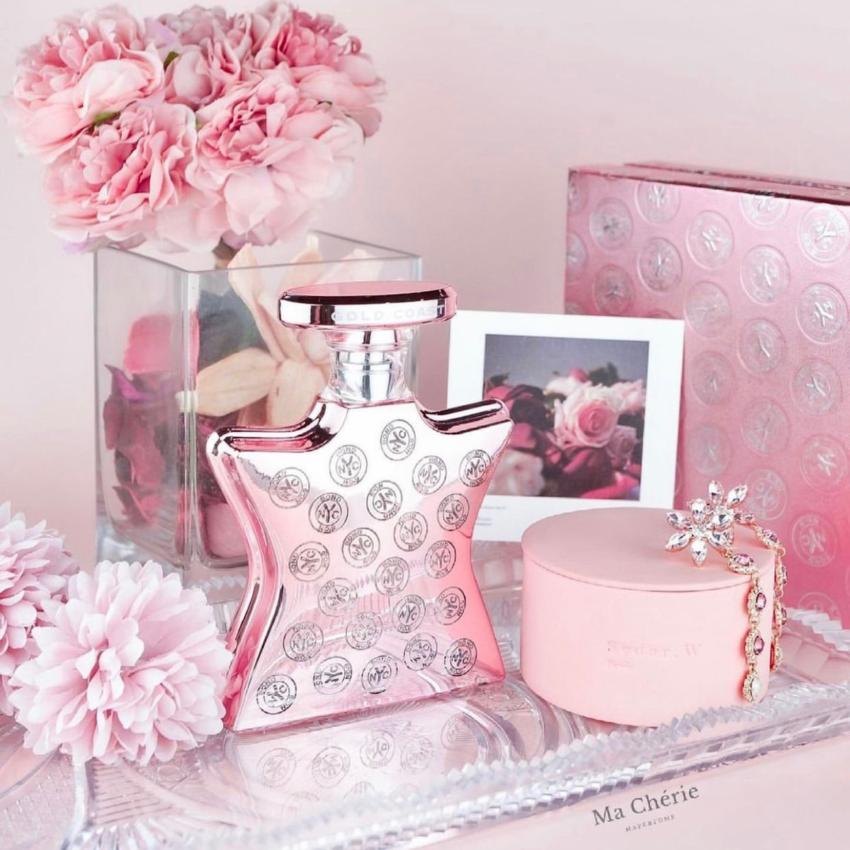 A pink perfume bottle with silver crystal-like circular embellishments, a round pink box, a small pink jewelry box with a rhinestone snowflake charm, and pink flowers arranged on a reflective tray against a soft pink background.