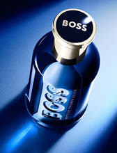 A bottle of HUGO BOSS Bottled Triumph Elitr cologne, featuring a dark blue glass bottle with a metallic gold cap and white lettering, set against a blue gradient background with dramatic lighting.