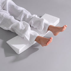 A person lying on a white foam heel lift leg pillow on a light gray studio floor, wearing loose white pants with both feet visible.