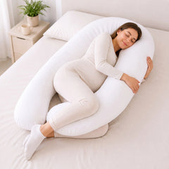 A woman in a cream-colored pajama set rests on a white U-shaped maternity pillow on a bed in a bright, neutral bedroom.