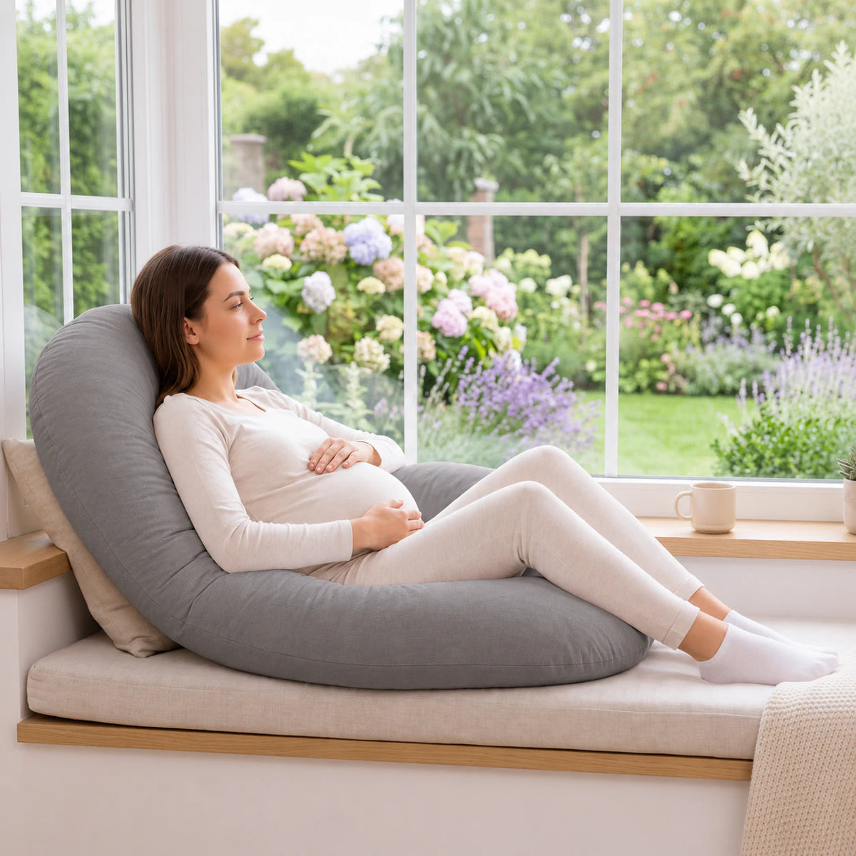 Woman reclining with a grey U-shaped pregnancy pillow on a window seat.